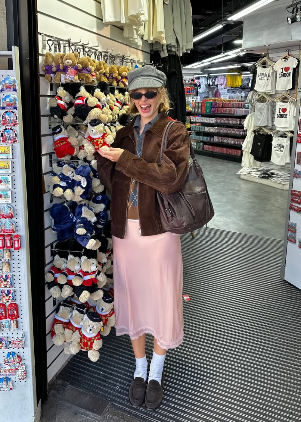 Rosa pastel em saia acetinada; modelo posa sorrindo com jaqueta marrom e boina cinza em loja de souvenirs londrina Texto: