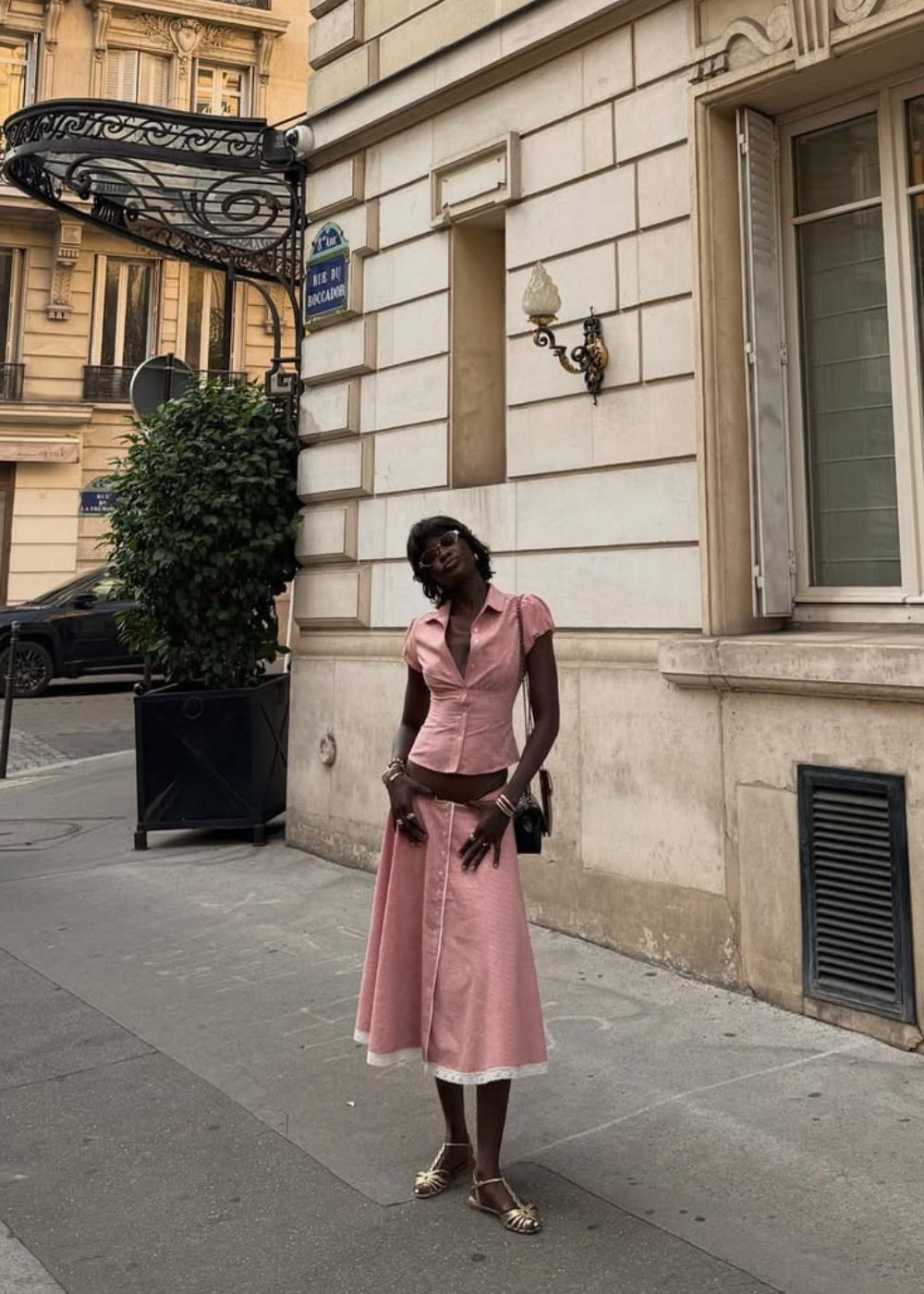 Rosa pastel em conjunto ajustado de blusa e saia midi; modelo posa elegante na calçada diante de fachada clássica.