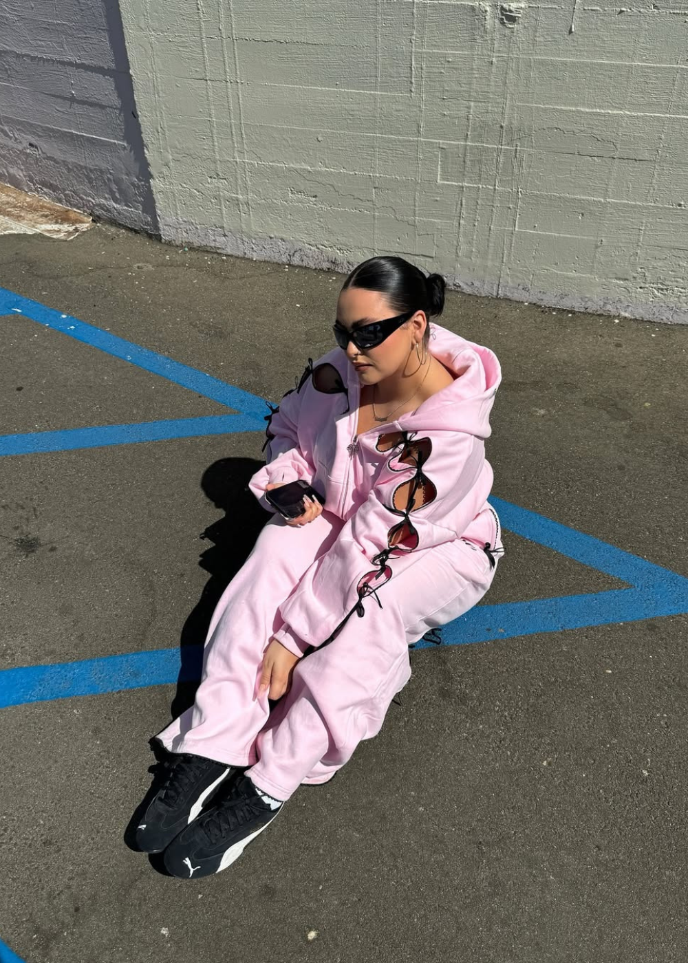 Rosa pastel em conjunto moletom amplo com recortes e amarrações; modelo posa sentada no asfalto sob sol urbano.