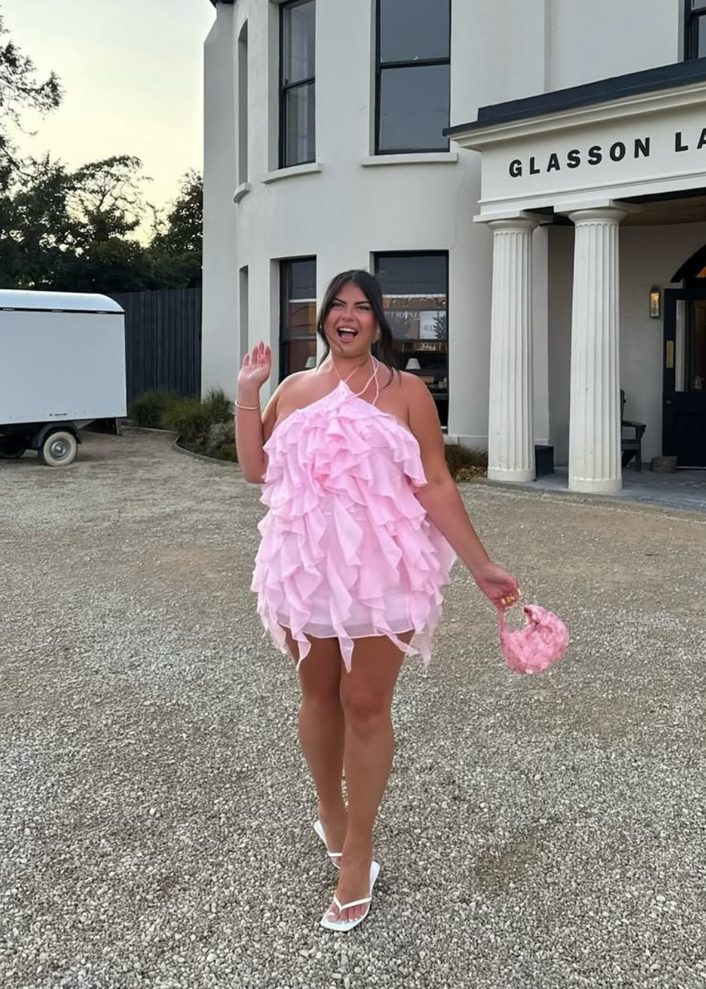 Modelo posa sorrindo com vestido curto de babados em rosa pastel, bolsa floral e salto branco. Texto: GLASSON LA