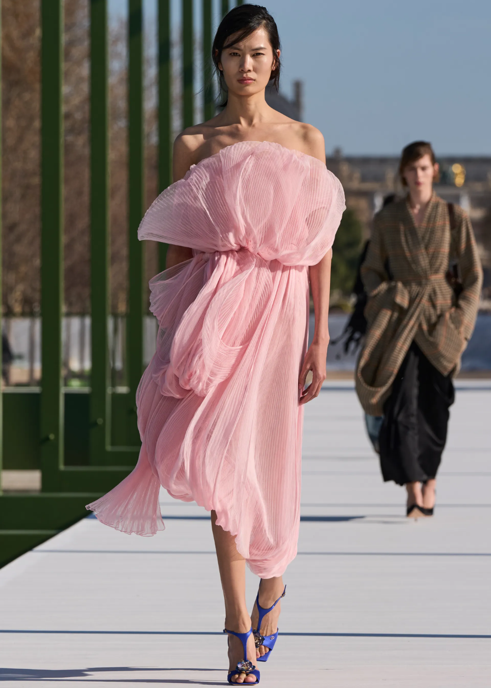 Rosa pastel em vestido plissado tomara que caia; modelo desfila ao ar livre com leveza, volume e sandálias azuis.