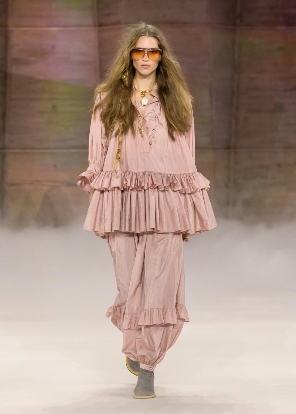 Modelo desfila em rosa pastel com túnica e calça amplas de babados, tecido leve, óculos âmbar e clima boho suave.