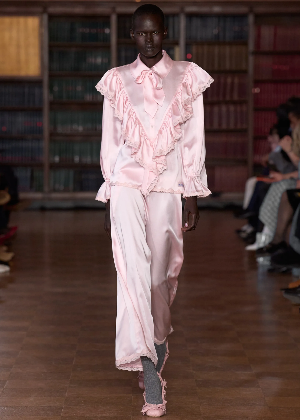 Rosa pastel em look de cetim com babados e renda; modelo desfila com elegância serena em passarela entre estantes.