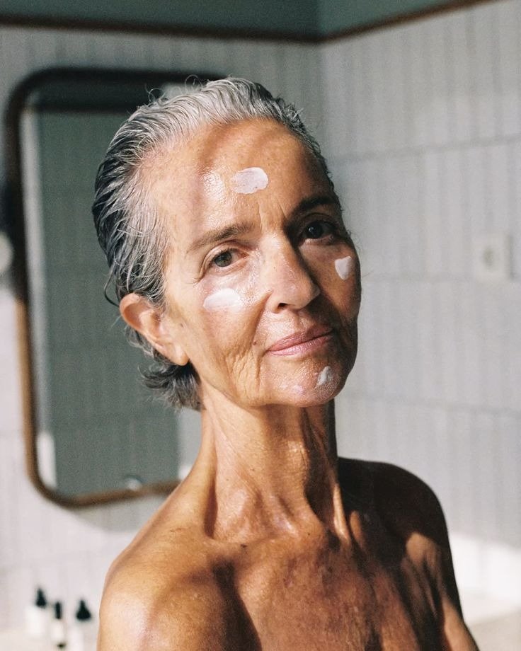 Melhor sérum para pele madura: mulher grisalha posa no banheiro com creme no rosto, luz suave e clima sereno