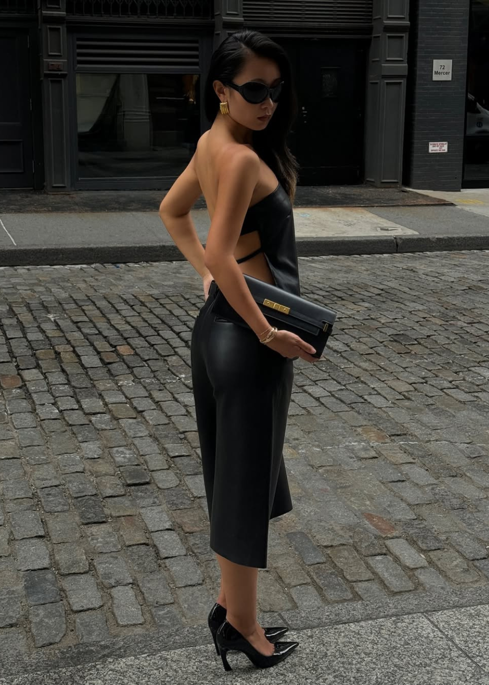 Modelo posa de macacão preto acetinado com recortes, clutch e scarpin de Salto vírgula em rua sóbria.