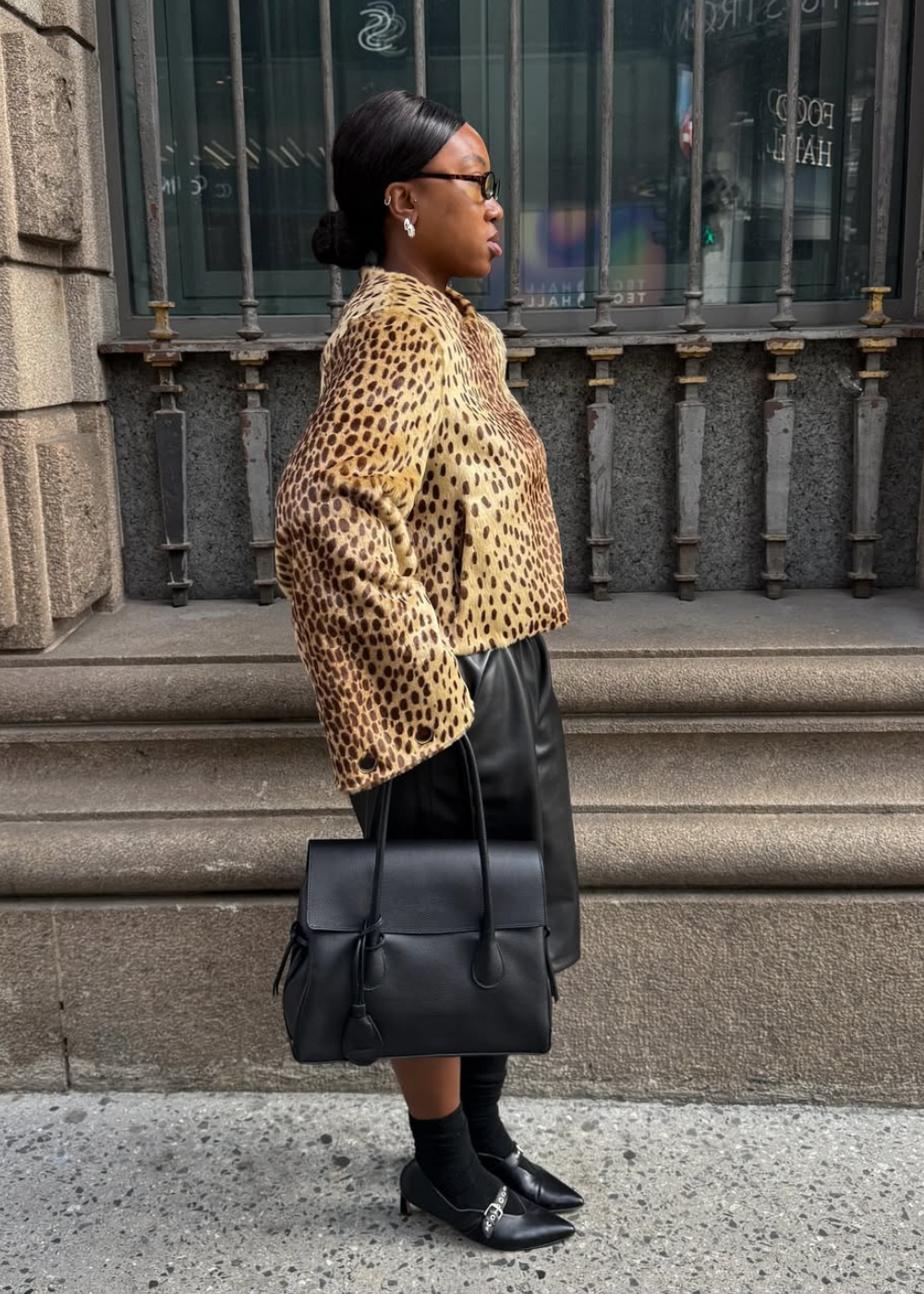 Salto vírgula em sapato preto com fivela; modelo posa de perfil com jaqueta animal print e bolsa estruturada, ar elegante.