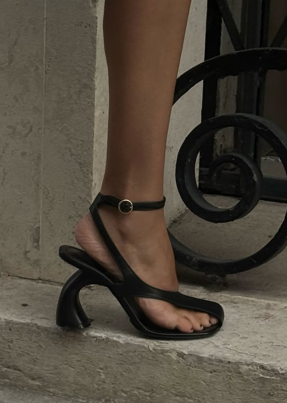 Sapatos arquitetônicos pretos de tiras e salto curvo escultural em pé na pedra criam um clima sofisticado e ousado