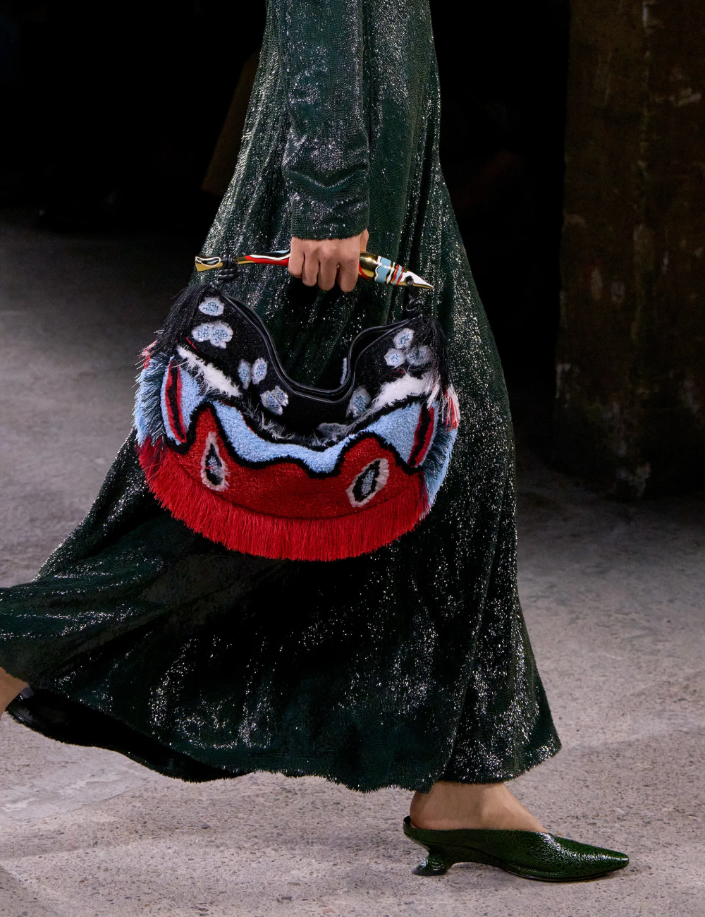 Modelo caminha com salto vírgula e vestido longo brilhante verde-escuro, segurando bolsa felpuda vermelha e azul.