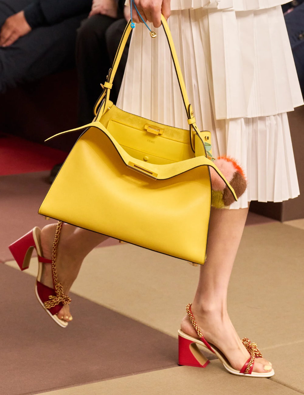 Salto vírgula em sandália vermelha com corrente dourada acompanha bolsa amarela e vestido plissado branco elegante.