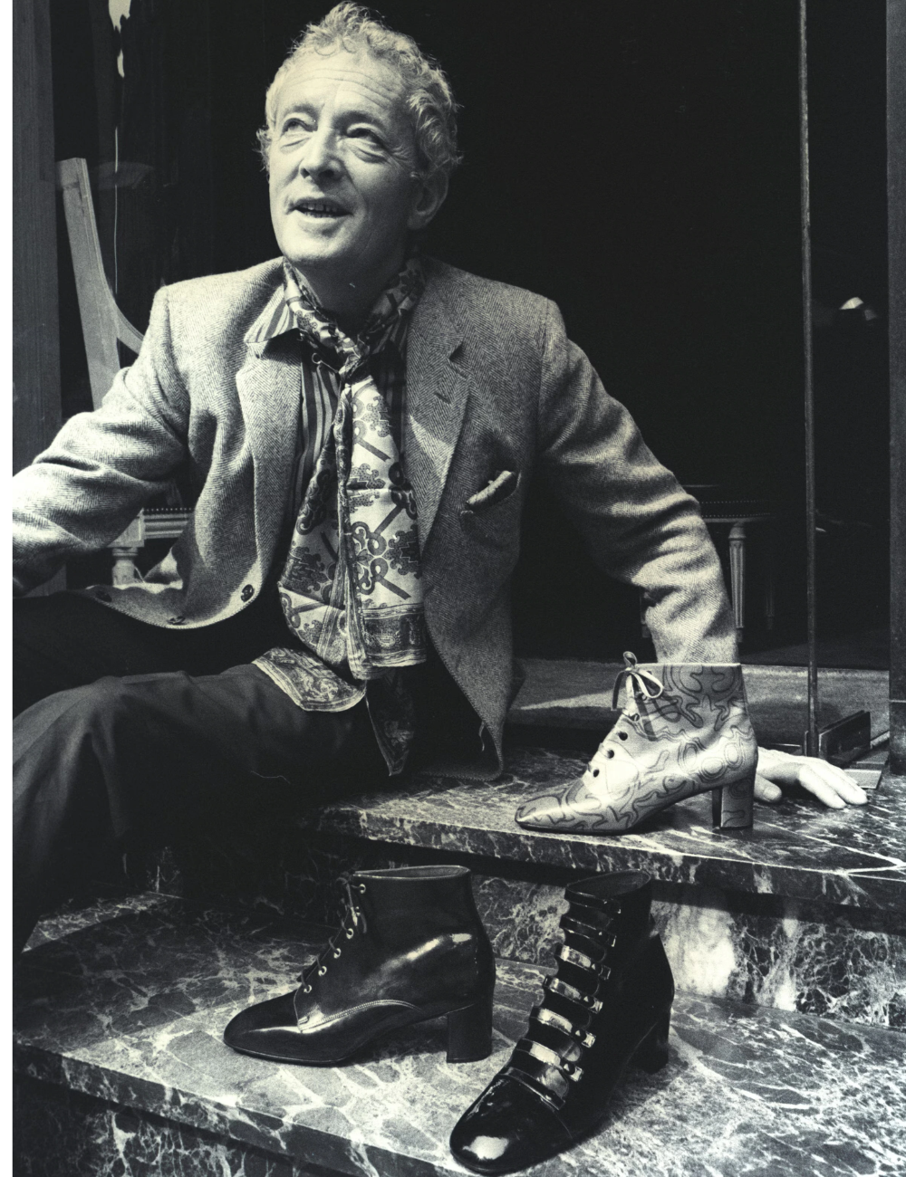 Salto vírgula em botas de couro e ankle boot estampada sobre mármore; homem posa de blazer cinza com elegância serena.