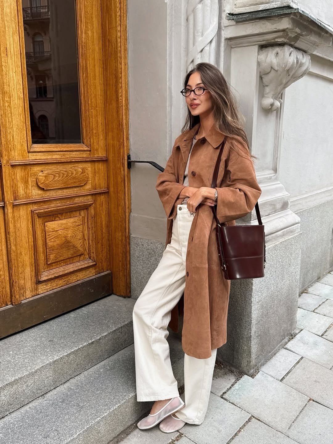Modelo posa com sapatilha de tela nude, trench marrom e calça off-white diante de fachada clássica, em clima elegante.