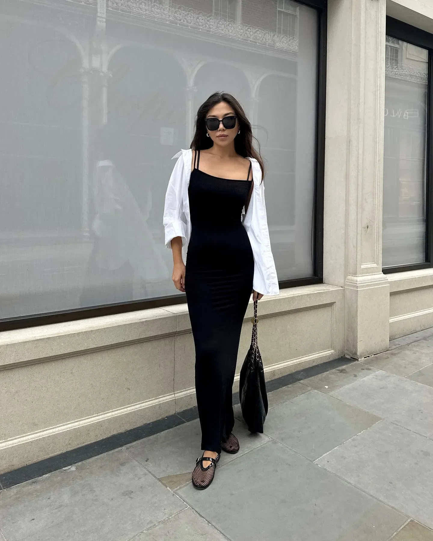 Modelo posa com vestido preto ajustado, camisa branca aberta e sapatilha de tela em look urbano elegante e sóbrio.