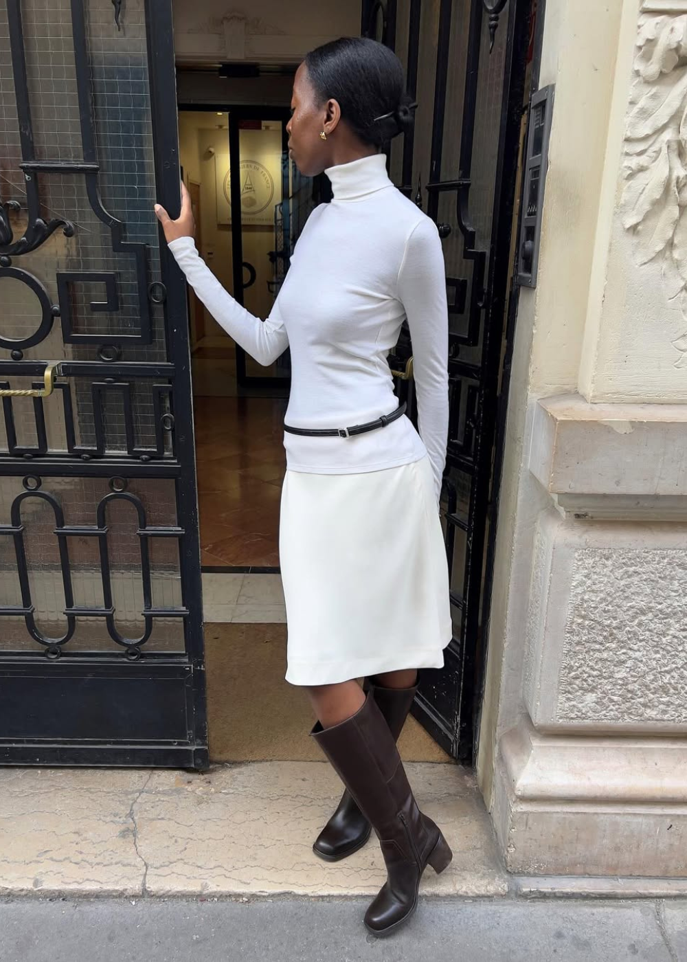 Sapatos do inverno 2026: modelo posa de perfil com bota marrom de cano alto, vestido branco e elegância serena.