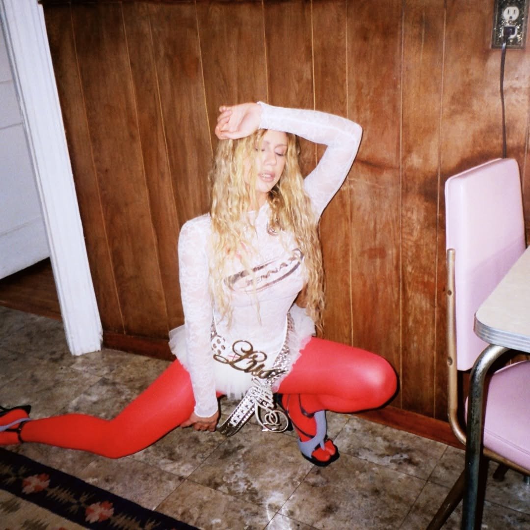 Slayyyter posa no chão com vestido branco justo e transparente, meia-calça vermelha e salto, em clima ousado retrô.