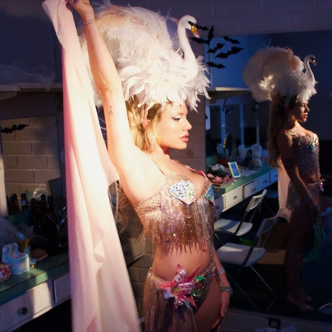 Slayyyter posa com look de franjas e brilho prateado, cocar de cisne branco e clima glamouroso nos bastidores.