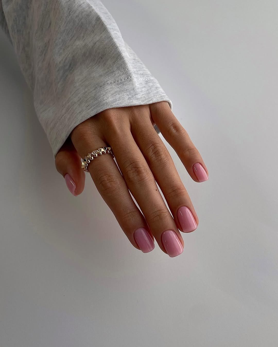 Unhas para madrinha de casamento em rosa-claro brilhante, mão posa com anel prateado e manga cinza em fundo clean.