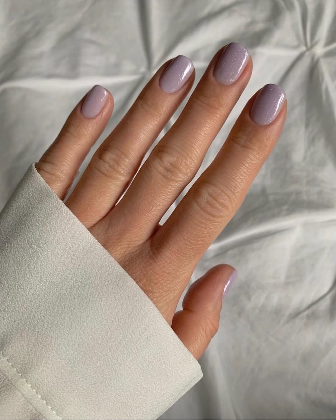 Unhas para madrinha de casamento em lilás claro cintilante, curtas e ovais, com manga branca e fundo de tecido suave.