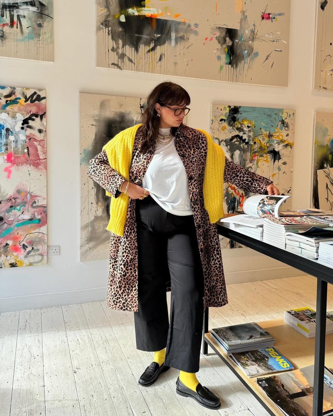 Modelos de calças de alfaiataria: modelo posa com calça preta ampla, casaco animal print e tricô amarelo em galeria