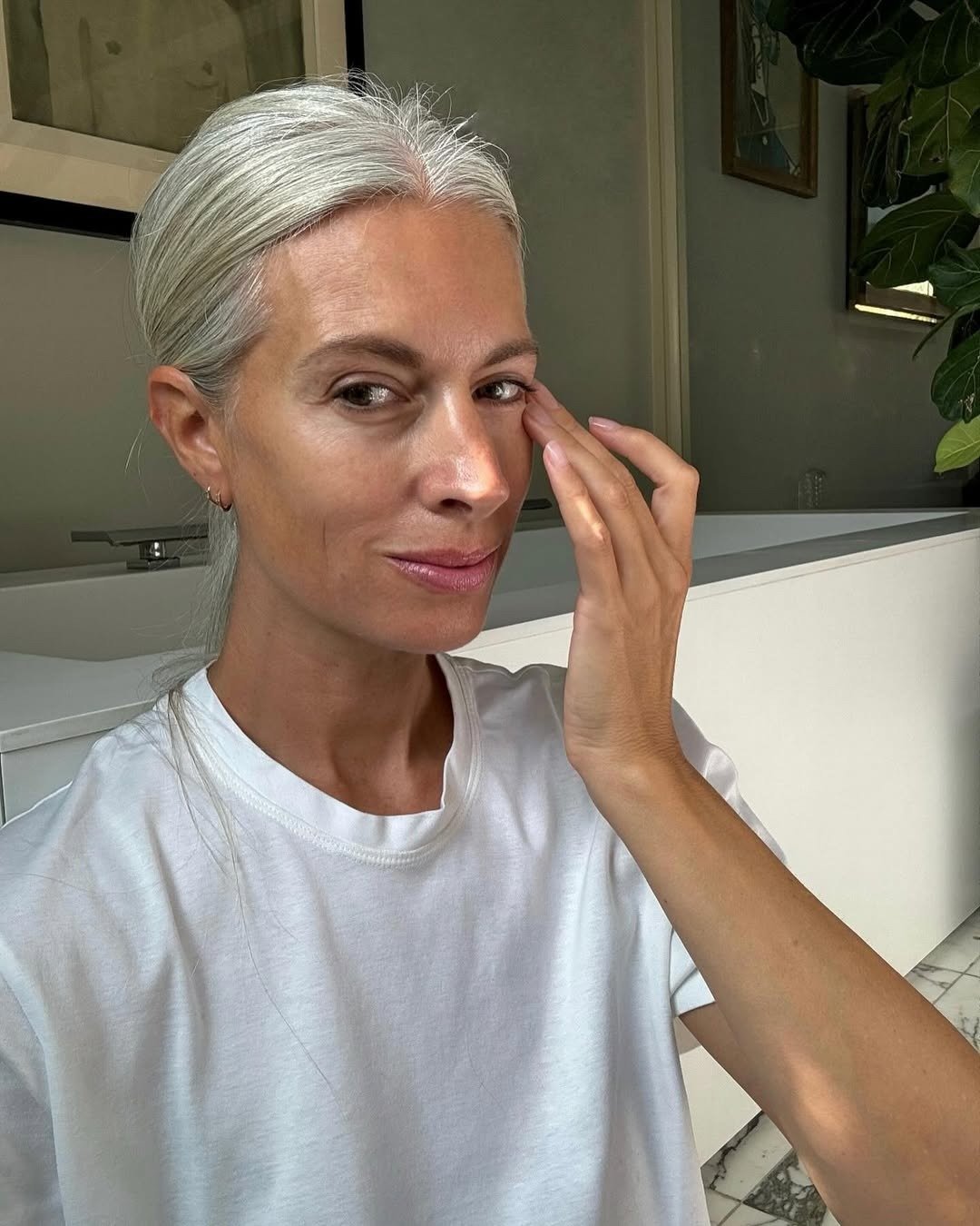 Melhor sérum para pele madura: mulher de camiseta branca aplica produto no rosto em banheiro claro, com expressão serena.