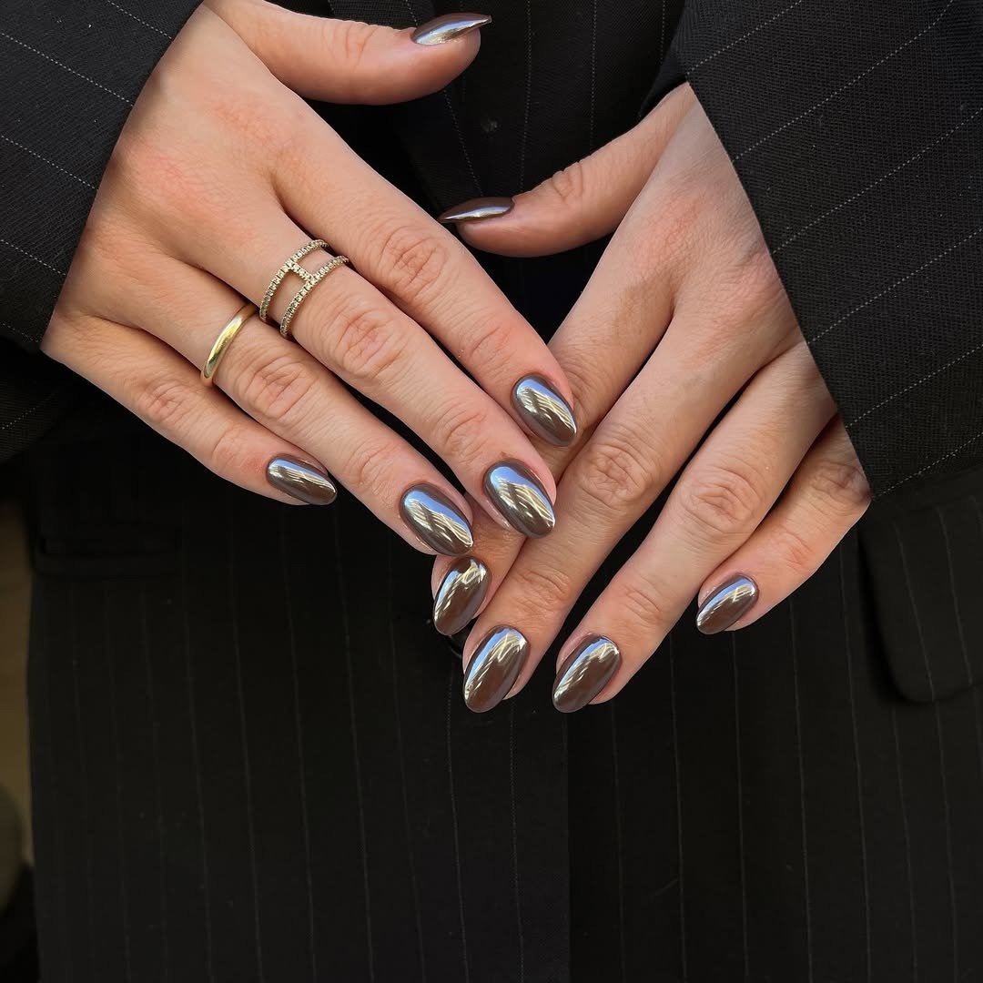 Unhas para madrinha de casamento em tom grafite espelhado, com anéis delicados e blazer risca de giz em pose elegante