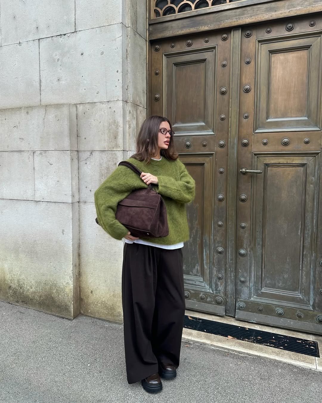 Looks estilosos para a variação de temperatura no outono: modelo posa com tricô verde, calça ampla e bolsa marrom.