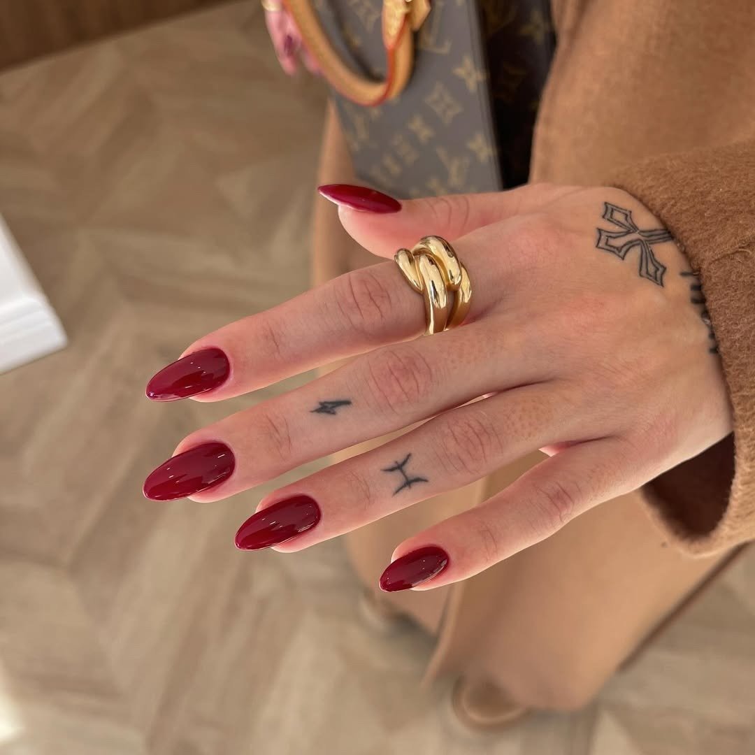 Unhas para madrinha de casamento em vinho brilhante e formato amendoado, com anel dourado e casaco camel elegante.
