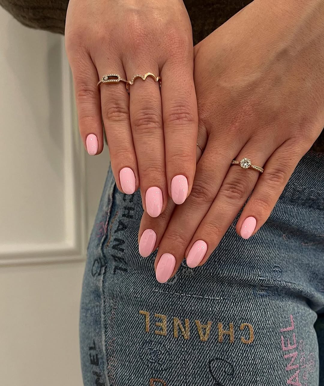 Unhas para madrinha de casamento em rosa-claro brilhante, formato oval, com anéis delicados sobre jeans.