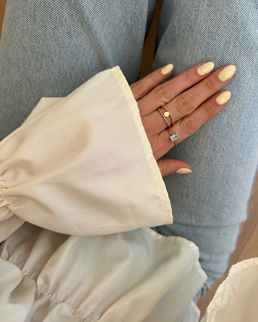 Unhas para madrinha de casamento em tom manteiga, formato amendoado, com blusa off-white fluida e anéis delicados dourados.