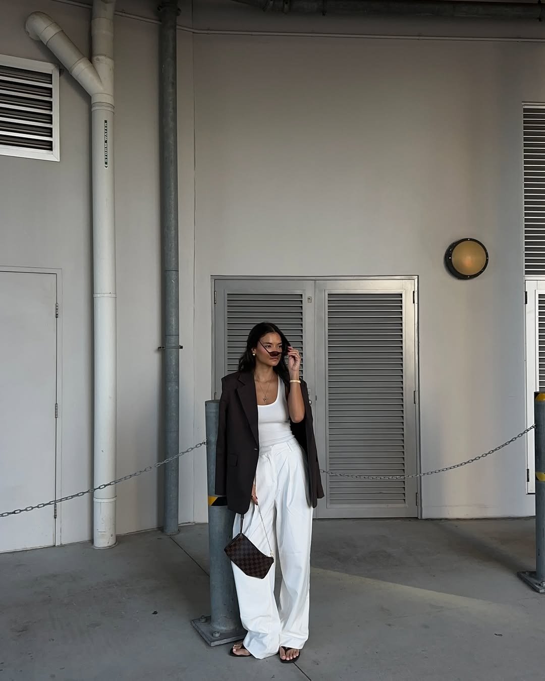 Modelo posa com calça branca ampla, regata e blazer marrom em cenário urbano neutro, com elegância casual.