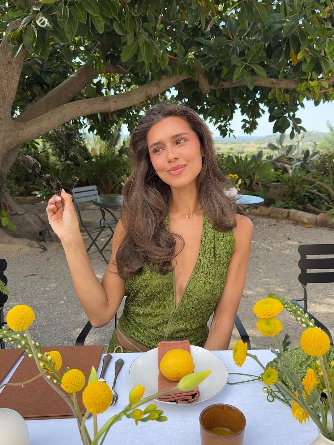 O que é sulfato no shampoo: modelo posa à mesa com vestido verde rendado, flores amarelas e clima leve ao ar livre