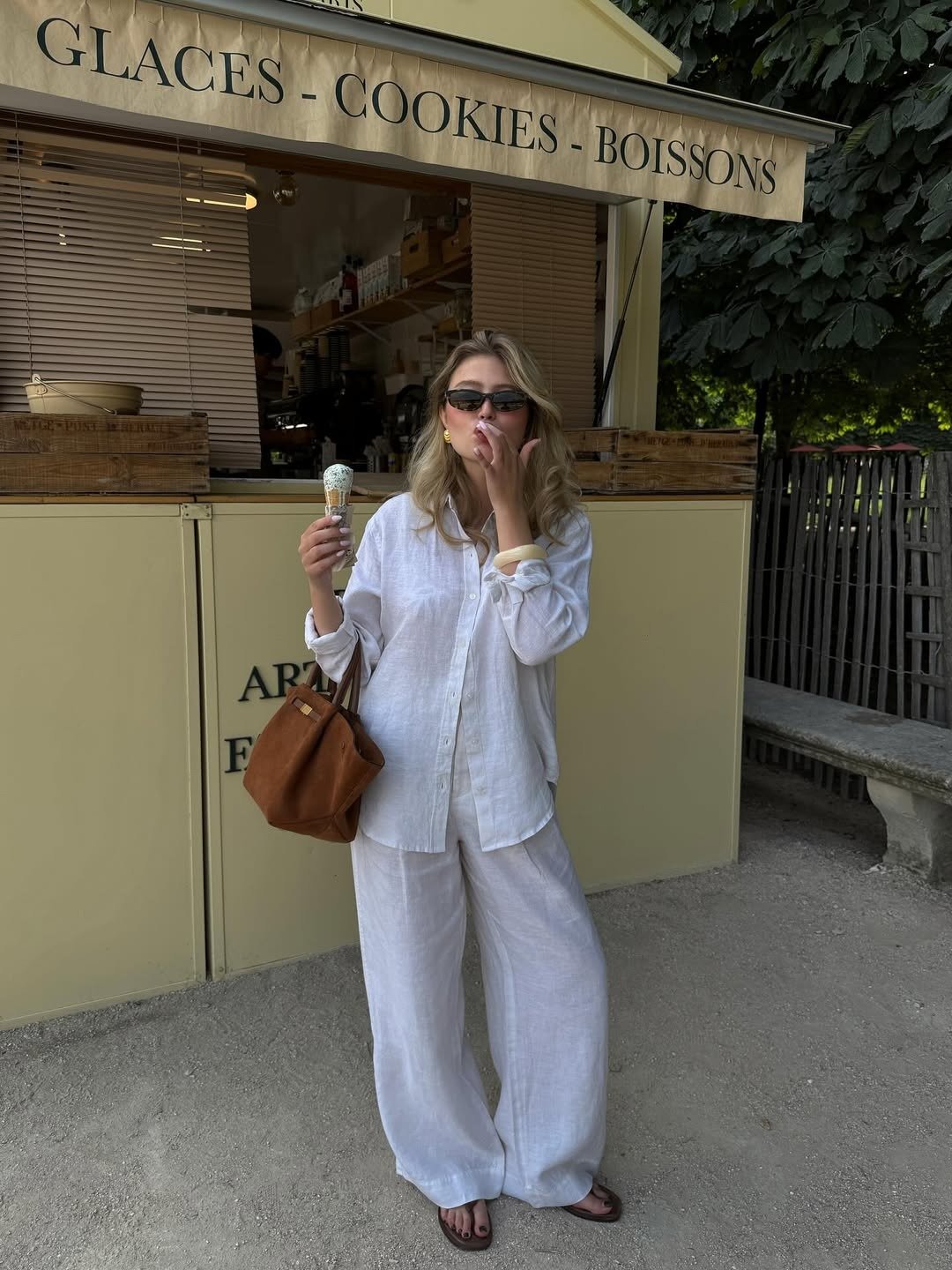 Modelo posa com calça branca pantalona e camisa de linho, sorvete na mão, clima leve. Texto: GLACES - COOKIES - BOISSONS