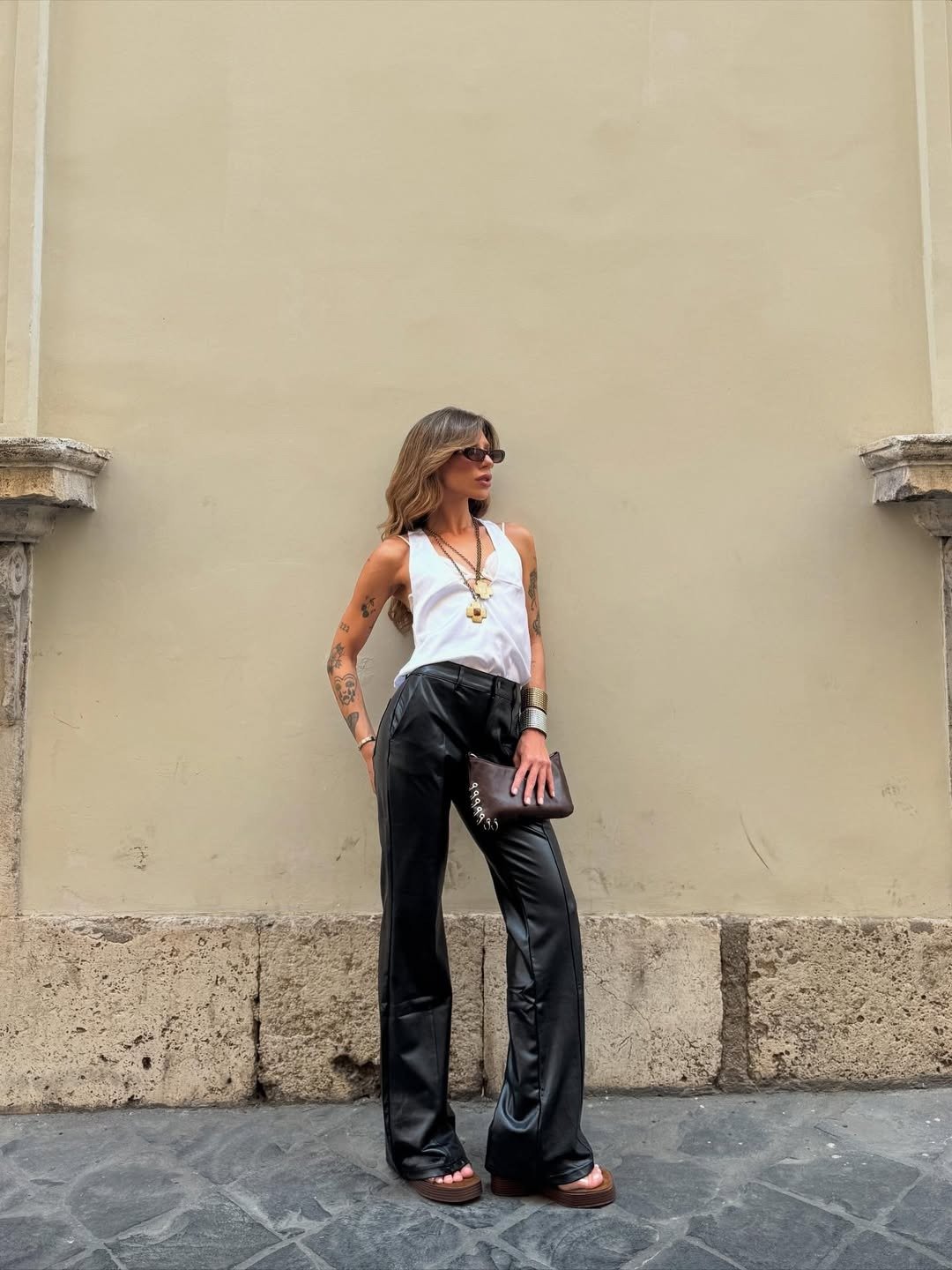 Looks com calça de couro: modelo posa com regata branca, pantalona preta, óculos e clutch marrom em clima urbano elegante.