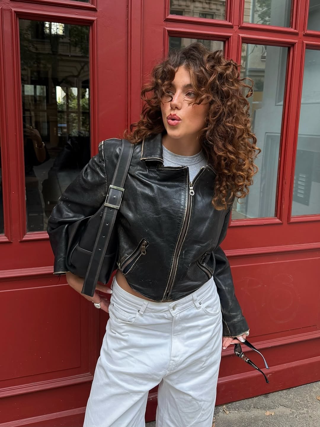 O que é sulfato no shampoo: modelo posa com jaqueta de couro preta, jeans branco e cachos marcados diante de porta vermelha.