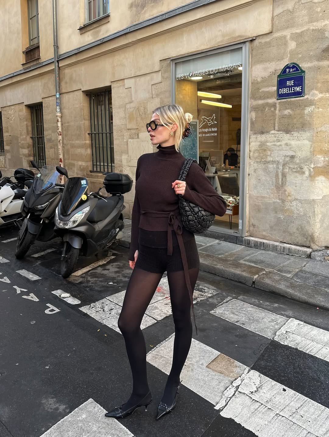 Looks estilosos para a variação de temperatura no outono: modelo posa com tricô marrom, meia-calça preta e bolsa matelassê.