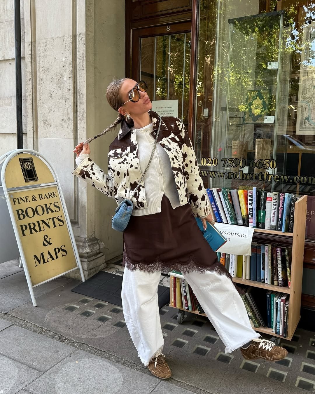 Modelo posa com calça branca ampla, jaqueta estampada e sobreposição marrom diante de livraria charmosa.
