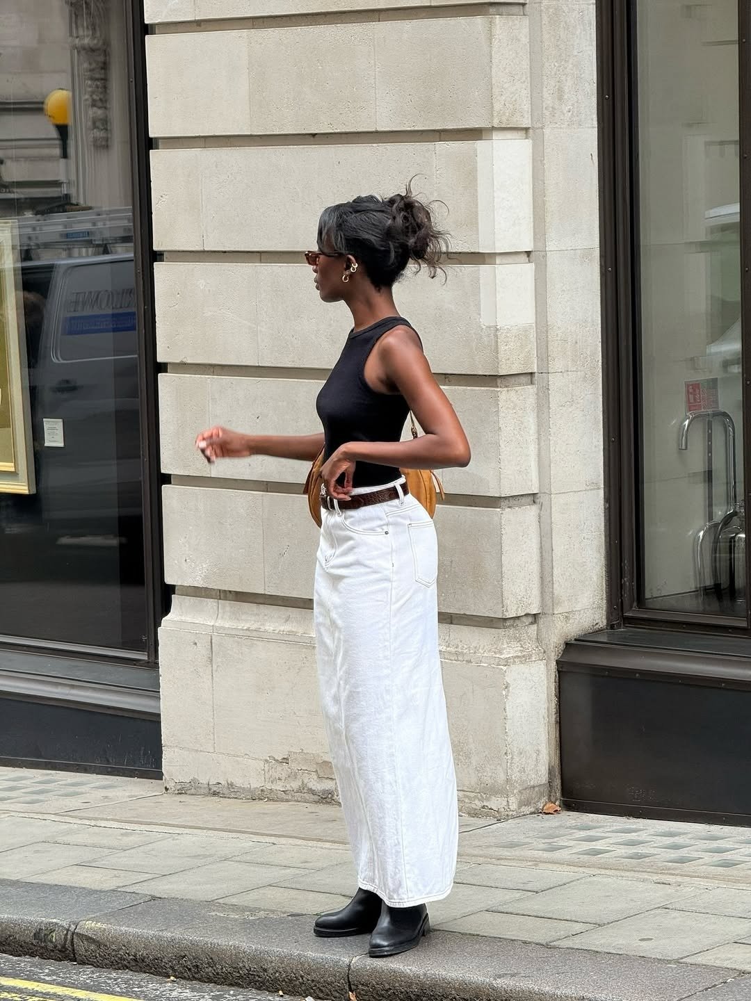 Modelo posa com calça branca longa de jeans, regata preta e bota preta em rua urbana, com elegância minimalista.