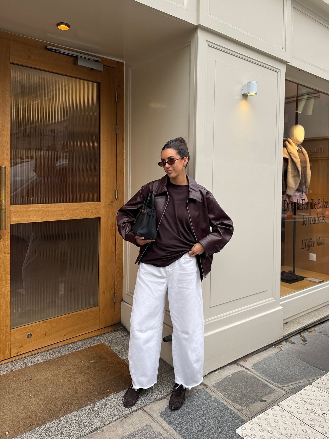 Modelo posa com calça branca ampla, jaqueta de couro vinho e bolsa preta na calçada, em clima urbano elegante.