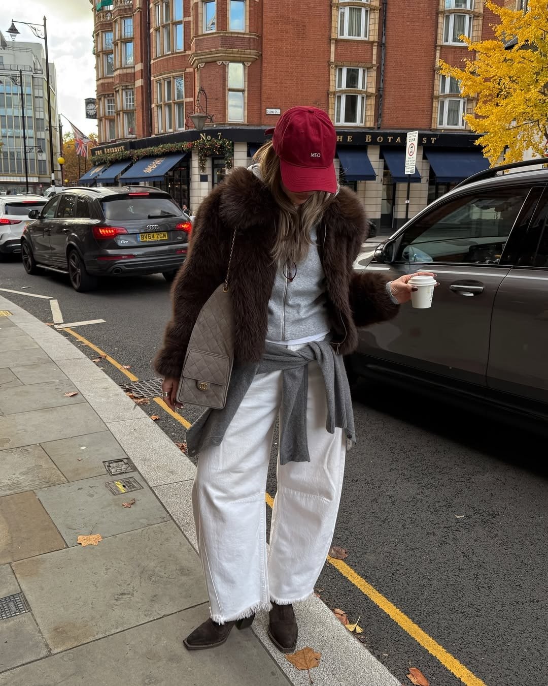 Modelo posa com calça branca ampla, jaqueta de pelo marrom e boné vinho em rua urbana de clima sofisticado.