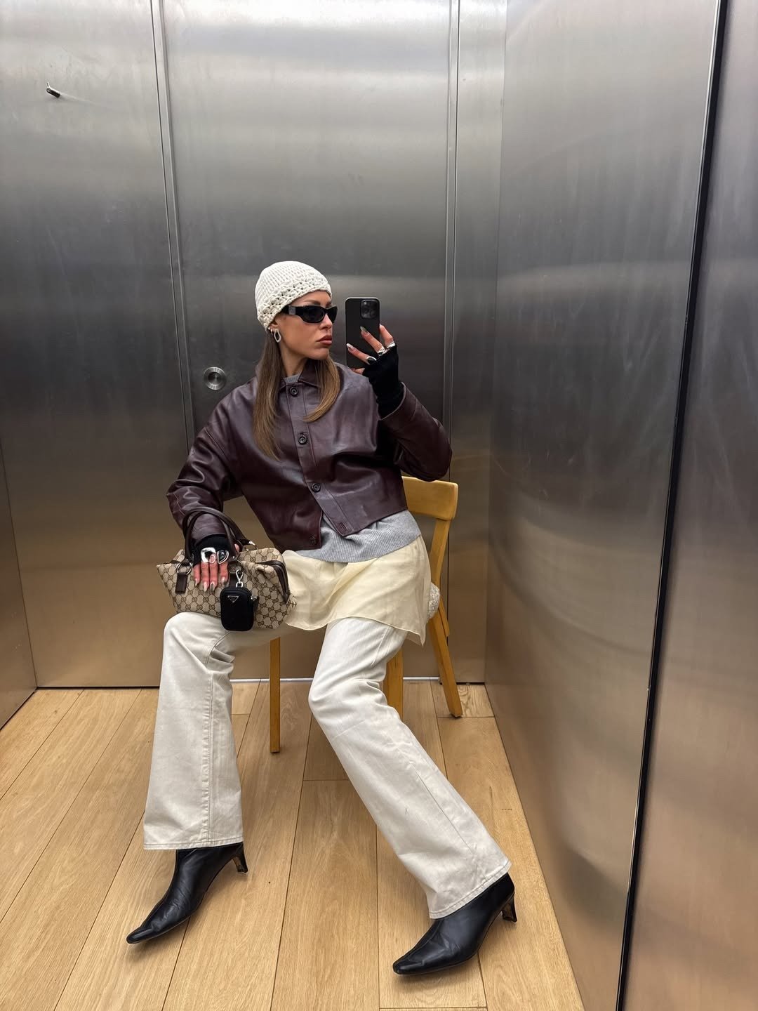 Modelo posa sentada com calça branca ampla, jaqueta marrom de couro, gorro de tricô e botas pretas em clima urbano