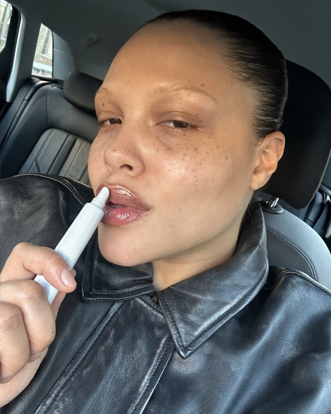 Modelo posa no carro com jaqueta de couro preta e gloss rosado, destacando cores de batom vermelho em clima urbano sóbrio