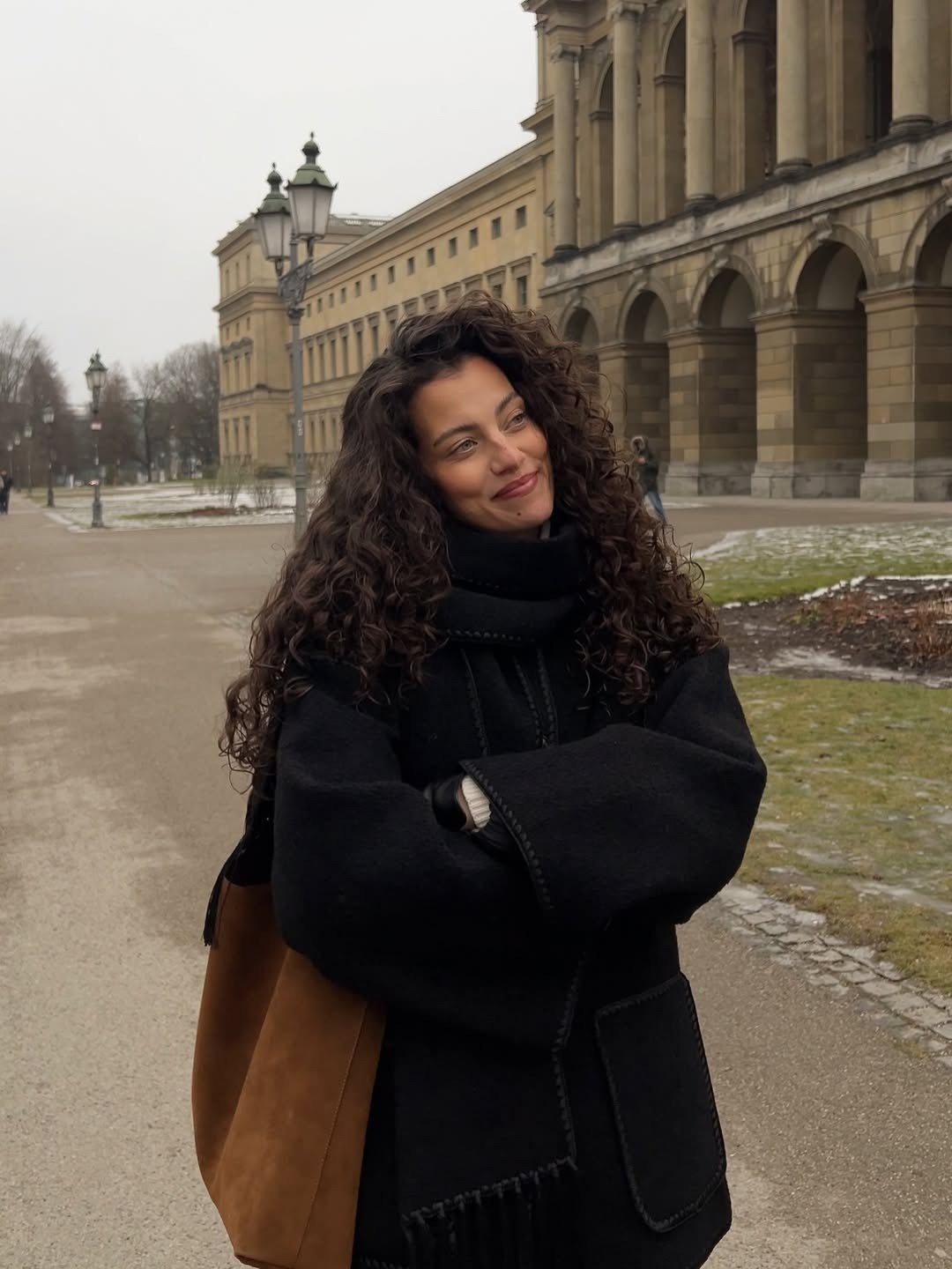 O que é sulfato no shampoo: modelo posa de casaco preto amplo e bolsa marrom diante de prédio clássico, em clima sereno.