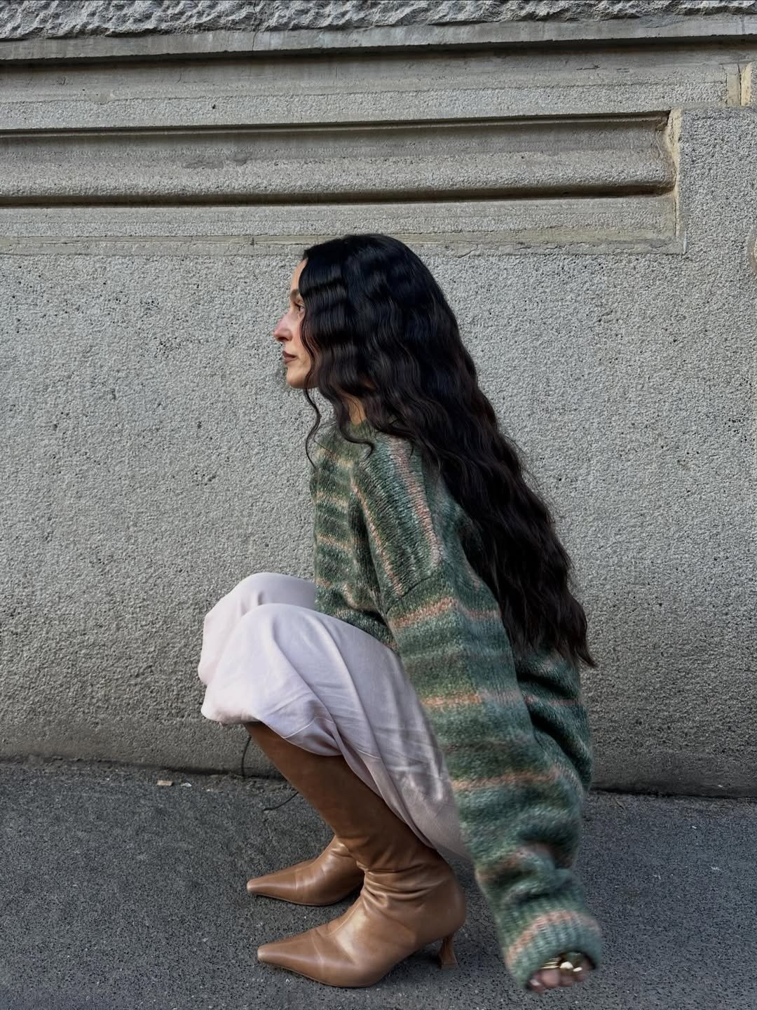 O que é sulfato no shampoo: modelo agacha com tricô verde mescla, saia branca e bota caramelo diante de muro cinza sereno
