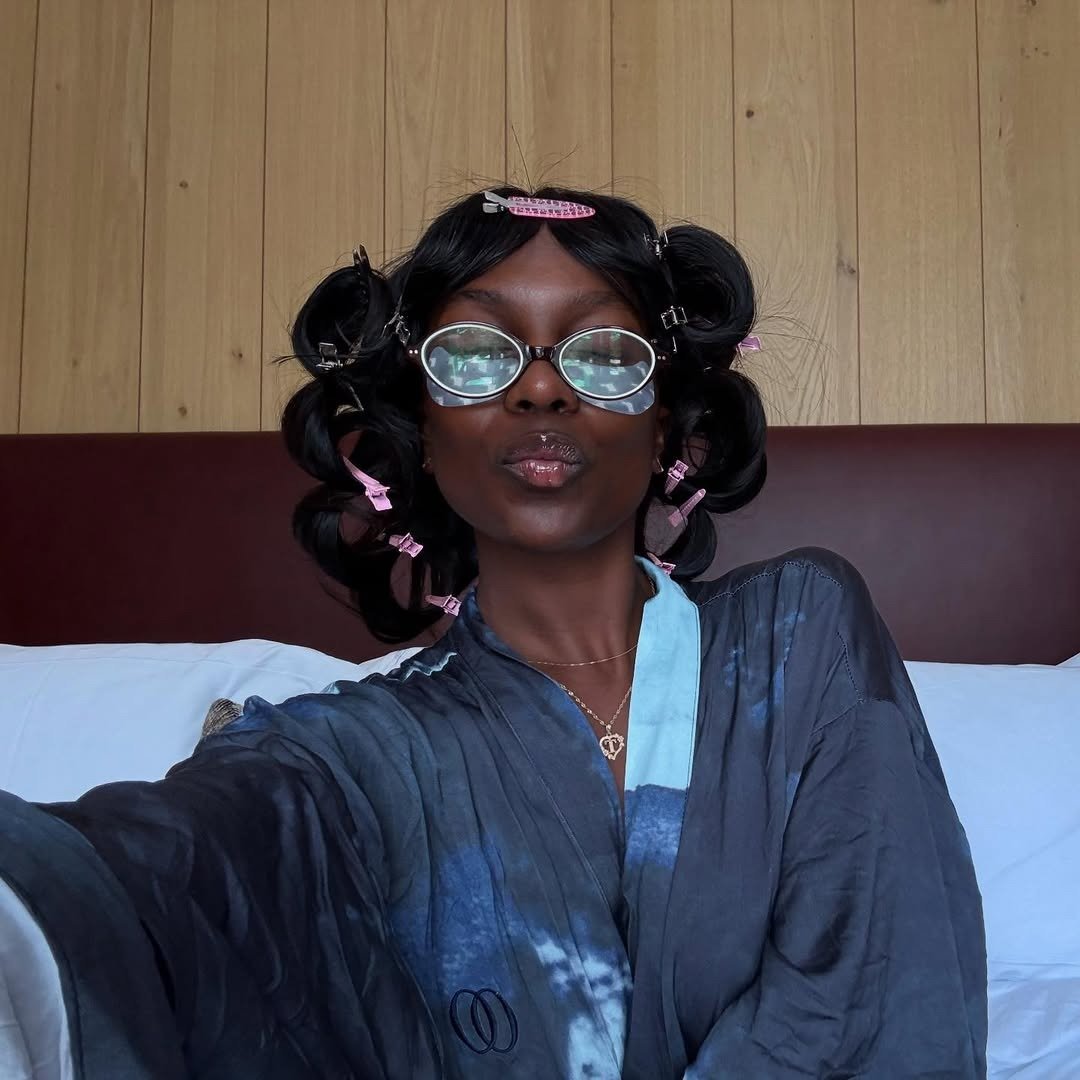 Benefícios da limpeza de pele: modelo posa de robe acetinado azul, óculos redondos e bobes rosa em clima leve e vaidoso