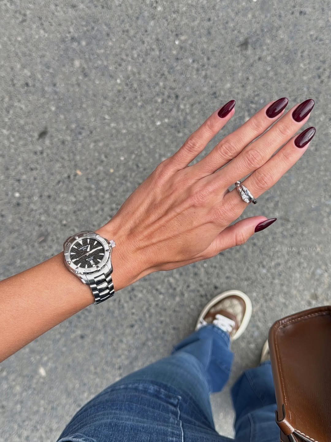 Unhas para madrinha de casamento em vinho brilhante, modelo posa com jeans azul, relógio prateado e bolsa marrom.
