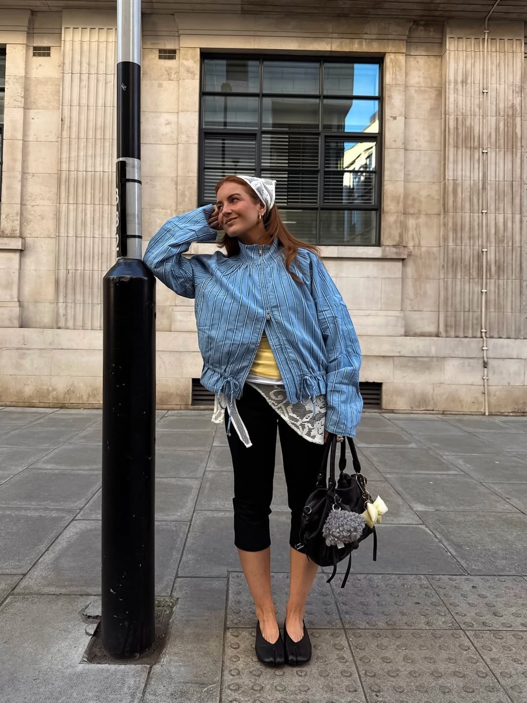Looks para explorar a criatividade: modelo posa com jaqueta azul listrada, lenço e bolsa preta em clima urbano leve.