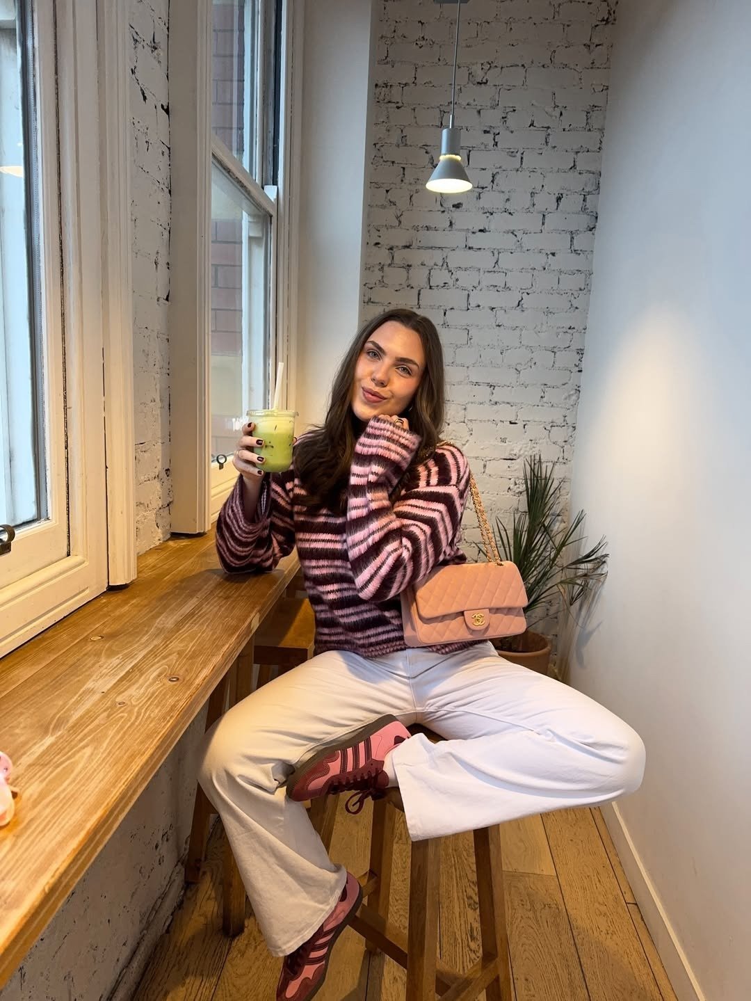Modelo posa com calça branca flare, tricô listrado rosa e vinho e bolsa matelassê em café acolhedor e leve.