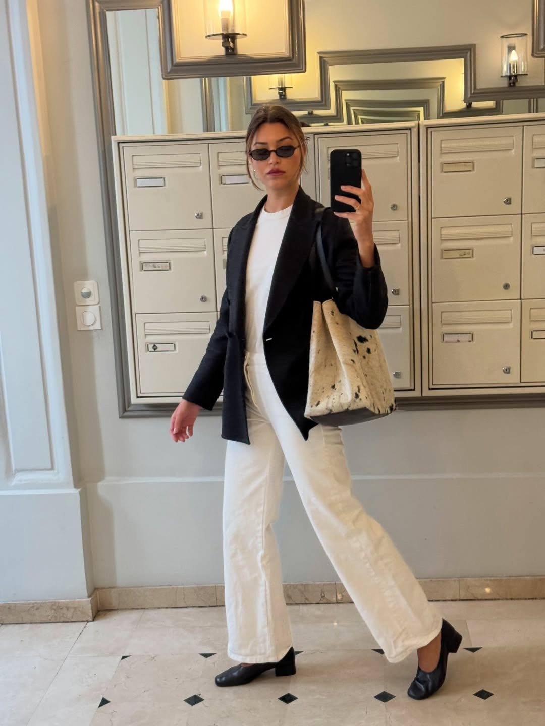 Modelo posa com calça branca reta, blazer preto e bolsa texturizada em hall elegante, com ar sofisticado.