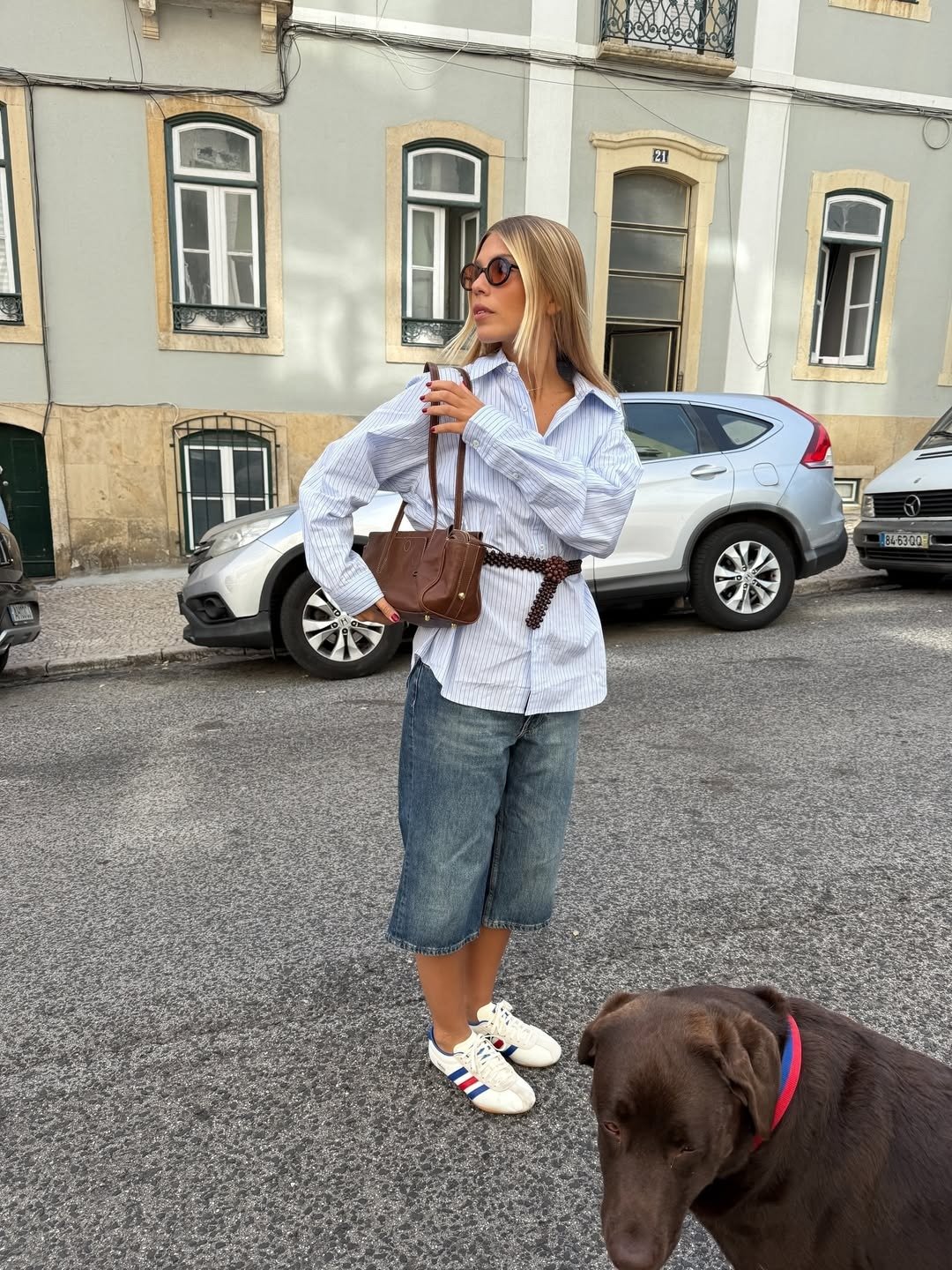 Looks estilosos para a variação de temperatura no outono: modelo posa com camisa listrada, bermuda jeans e bolsa marrom.