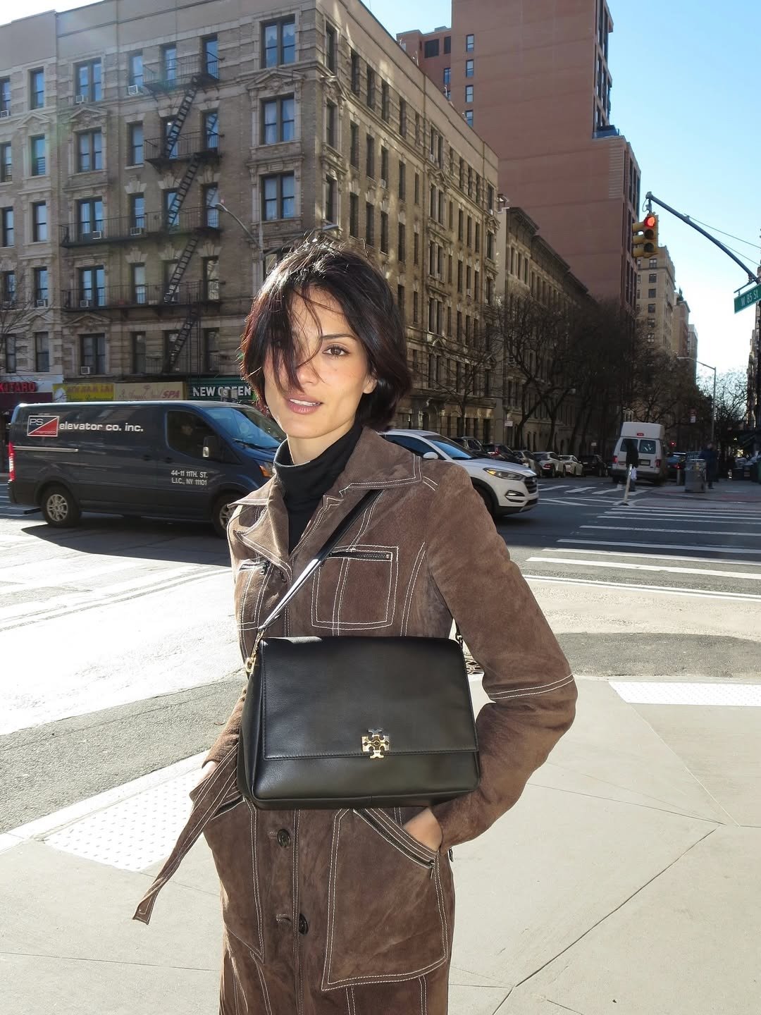 O que é sulfato no shampoo: modelo posa de casaco marrom pespontado, gola preta e bolsa estruturada em rua ensolarada.