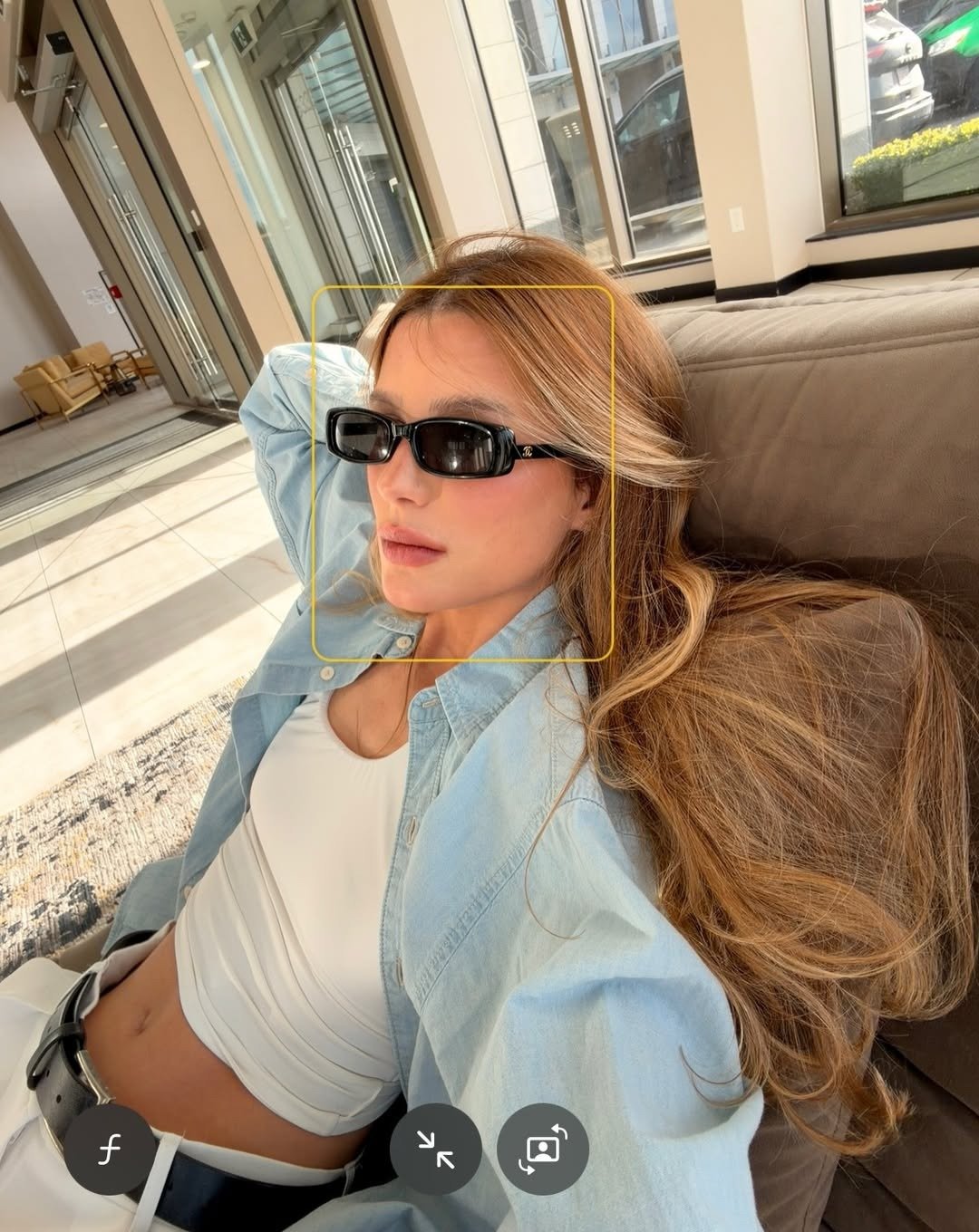 O que é sulfato no shampoo: modelo relaxa no sofá com camisa azul, top branco, óculos pretos e luz dourada elegante